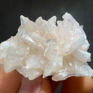 May include: A cluster of pale pink crystal formations. The crystals are elongated and pointed, creating a textured, spiky appearance. The mineral specimen has a soft, translucent quality, with subtle variations in color. The background is a dark gray.