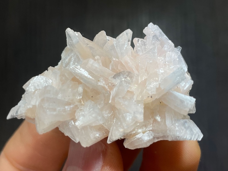 May include: A cluster of pale pink crystal formations. The crystals are elongated and pointed, creating a textured, spiky appearance. The mineral specimen has a soft, translucent quality, with subtle variations in color. The background is a dark gray.