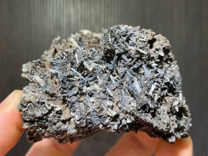 May include: A close-up of a mineral specimen featuring a cluster of dark, needle-like crystals. The crystals are primarily black and gray, with some lighter, translucent areas. The matrix is a mix of brown and gray, creating a textured appearance.