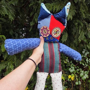 May include: A handmade fabric doll with a patchwork head featuring red, blue, and patterned fabrics. The doll has blue patterned arms, a striped torso, and white legs with a repeating red floral design. The doll is held against a backdrop of green foliage.