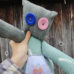 May include: A handmade plush toy in the shape of a person with green fabric, blue and pink buttons for eyes, and a fairy applique on the front.