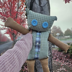 May include: A handmade plush toy made from repurposed fabric. The toy is shaped like a person with a plaid shirt, brown pants, and blue button eyes. The toy is hanging from a tree branch.