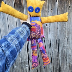 Pode incluir: Uma boneca de tecido amarelo feita &agrave; m&atilde;o com olhos de bot&atilde;o azul e pernas com padr&atilde;o colorido. A boneca est&aacute; em pose com os bra&ccedil;os estendidos.