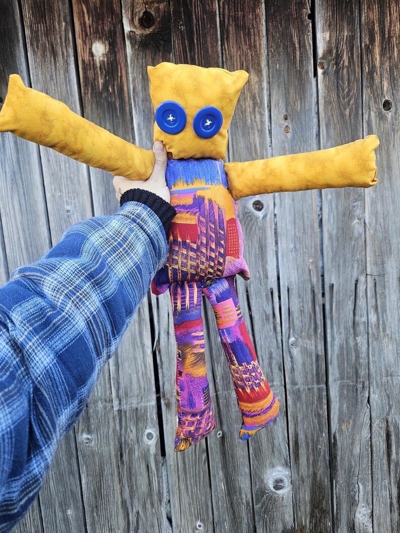 Pode incluir: Uma boneca de tecido amarelo feita &agrave; m&atilde;o com olhos de bot&atilde;o azul e pernas com padr&atilde;o colorido. A boneca est&aacute; em pose com os bra&ccedil;os estendidos.