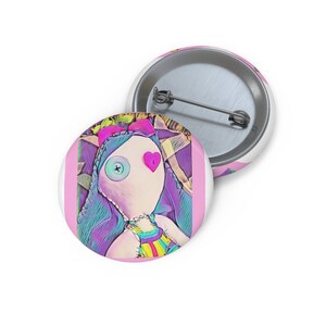 Könnte beinhalten: Zwei runde, weiße Knöpfe mit einem Cartoon-Puppe-Design. Die Puppe hat ein genähtes Gesicht, ein Knopfauge und ein herzförmiges Auge. Die Puppe hat blaue Haare und trägt ein regenbogenfarbenes Kleid. Die Knöpfe befinden sich auf einem rosa Hintergrund.