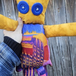 Pode incluir: Um brinquedo de pel&uacute;cia feito &agrave; m&atilde;o com uma cabe&ccedil;a amarela, olhos de bot&atilde;o azul e tecido estampado colorido para o corpo e os membros.