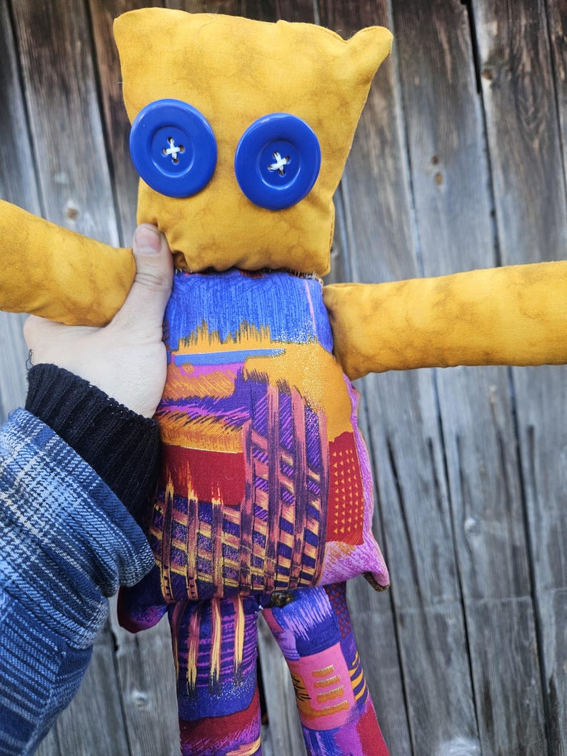 Pode incluir: Um brinquedo de pel&uacute;cia feito &agrave; m&atilde;o com uma cabe&ccedil;a amarela, olhos de bot&atilde;o azul e tecido estampado colorido para o corpo e os membros.