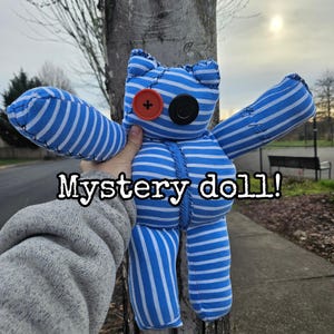 May include: A blue and white striped stuffed toy with a red button eye and a black button eye. The toy is standing on a tree trunk and the text "Mystery doll!" is written below it.