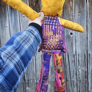 Pode incluir: Um brinquedo de pel&uacute;cia feito &agrave; m&atilde;o com a forma de uma pessoa com cabe&ccedil;a amarela, cal&ccedil;as com padr&atilde;o roxo e laranja e uma camisa com padr&atilde;o roxo e laranja. O brinquedo &eacute; segurado por uma m&atilde;o na frente de um fundo de madeira.