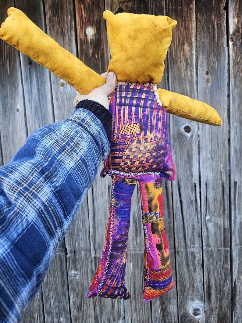 Pode incluir: Um brinquedo de pel&uacute;cia feito &agrave; m&atilde;o com a forma de uma pessoa com cabe&ccedil;a amarela, cal&ccedil;as com padr&atilde;o roxo e laranja e uma camisa com padr&atilde;o roxo e laranja. O brinquedo &eacute; segurado por uma m&atilde;o na frente de um fundo de madeira.