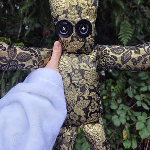 May include: A handmade doll with a black and gold floral pattern. The doll has large black button eyes and outstretched arms. The doll is made of fabric and has a whimsical, folk art aesthetic.
