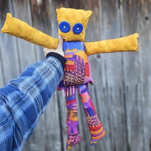 Pode incluir: Uma boneca de tecido amarela com olhos de bot&atilde;o azul e pernas de tecido com padr&atilde;o colorido. A boneca est&aacute; sendo segurada por uma m&atilde;o que est&aacute; usando uma camisa xadrez azul.