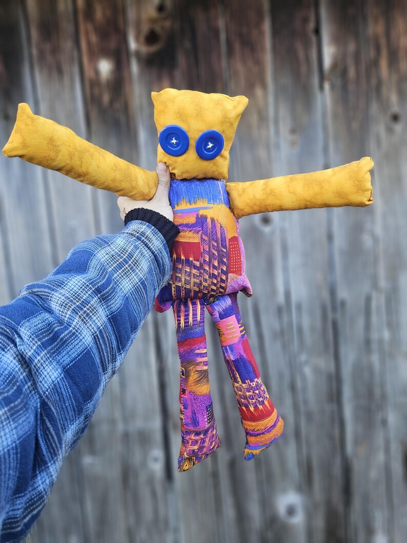 Pode incluir: Uma boneca de tecido amarela com olhos de bot&atilde;o azul e pernas de tecido com padr&atilde;o colorido. A boneca est&aacute; sendo segurada por uma m&atilde;o que est&aacute; usando uma camisa xadrez azul.