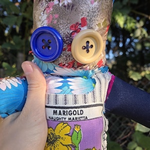 May include: Handmade fabric doll with a floral patterned head and body. The doll has large blue and tan button eyes. The doll's torso features a label that reads "MARIGOLD NAUGHTY MARIETTA" with a yellow flower illustration.