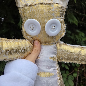 May include: A handmade owl doll with large white button eyes. The owl is crafted from gold and white patterned fabric, with a white body and gold wings. The doll is held by a person wearing a white robe.