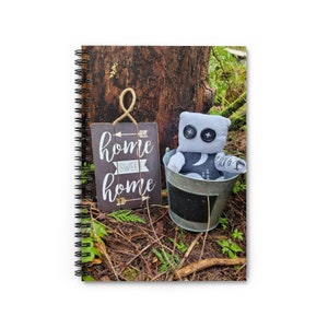 May include: A grey and white fabric robot toy sits in a metal bucket with a black chalkboard bottom. The bucket is on a bed of brown leaves and twigs. A wooden sign with the text "home sweet home" is in the background.