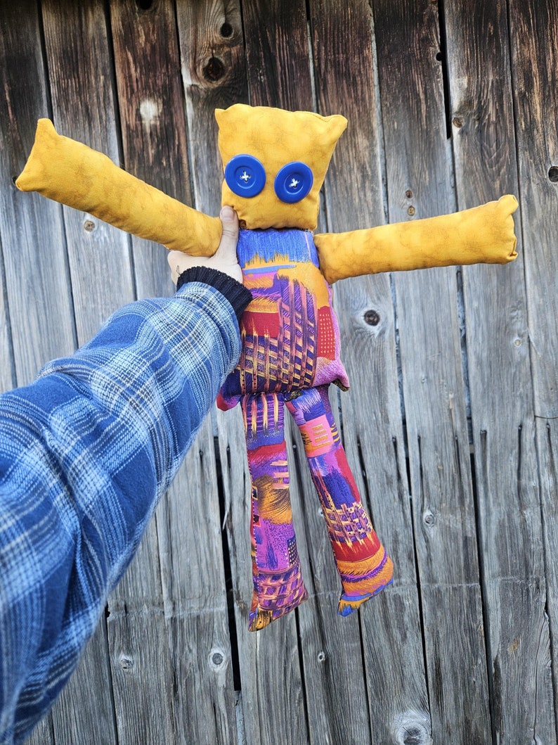 Pode incluir: Uma boneca de pano feita &agrave; m&atilde;o com bra&ccedil;os e pernas amarelos, um torso estampado em roxo, vermelho e azul e olhos de bot&atilde;o azuis.