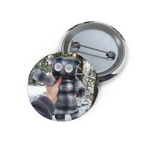 May include: Two round pin badges. One shows a handmade doll with button eyes and a plaid shirt. The other badge has a snowy forest scene. The doll is held in front of a snowy forest background.