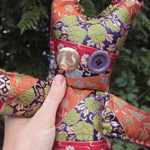May include: A handmade fabric doll with a patchwork design. The doll features orange, red, and navy blue floral patterns. It has a gold button and a purple button. The doll is held in a hand.