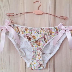 May include: White underwear with a floral and bunny print, trimmed with white lace. The sides are tied with light pink satin bows. The garment is hanging on a rose gold hanger.