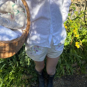 Puede incluir: Una persona con una camisa de lino blanca y pantalones cortos beige con bordados florales. La persona sostiene una cesta de mimbre llena de ropa de cama blanca. Lleva botas de goma negras y está en un jardín.