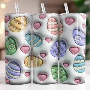 Könnte beinhalten: Drei weiße Edelstahlbecher mit einem pastellfarbenen Osterei-Design. Die Becher zeigen eine Vielzahl von pastellfarbenen Eiern mit verschiedenen Mustern, darunter Streifen, Punkte und Wirbel. Die Eier sind von rosa Herzen umgeben.
