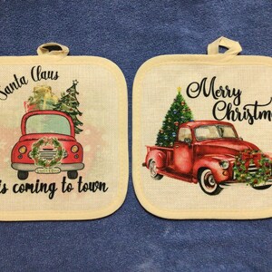 May include: Two cream-colored pot holders with red trucks and Christmas-themed designs. The first pot holder has a red truck with a Christmas tree and the text "Santa Claus is coming to town". The second pot holder has a red truck with a Christmas tree and the text "Merry Christmas".