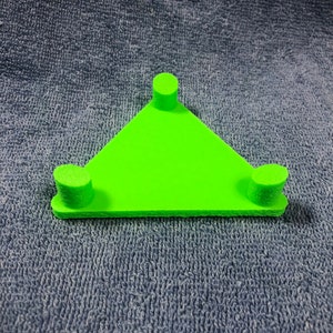 May include: A bright green, triangular object with three cylindrical supports. The object is made of a solid material and sits on a textured blue surface. The supports are evenly spaced along the edges of the triangle.