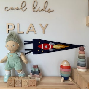 May include: A wooden sign with the words "cool kids play" in white lettering. A blue felt pennant with a red and white rocket ship and gold stars hangs on the wall. A doll with blonde hair and a blue outfit stands on a wooden shelf with wooden blocks that spell "zoom".