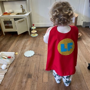 May include: A child wearing a red superhero cape with a gold circle and the letters "LF" in blue. The child has curly blonde hair and is standing on a wooden floor.