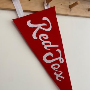 May include: A red pennant with the words "Red Fox" in white script. The triangular pennant hangs from a white ribbon. The background features a white wall and a wooden rack.