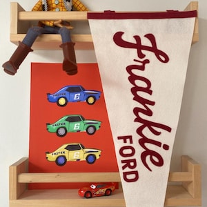 May include: A wooden shelf displays a Woody doll, a red poster with toy cars, a pennant with "Frankie FORD" in burgundy, and a toy car. The cars are blue, green, and yellow with the word "FASTER" and the number "6".