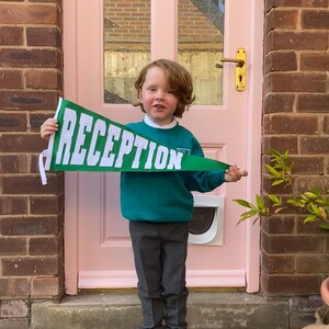 May include: A young child wearing a green jumper and grey trousers holds a green pennant with the word "RECEPTION" in white letters. The child is standing in front of a pink door.