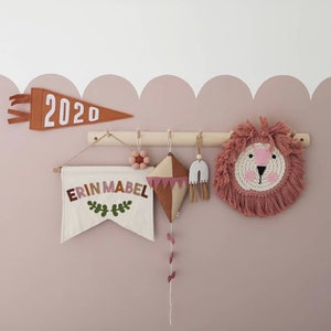 May include: A wooden wall rack displaying a collection of hanging nursery decor including a rust-coloured pennant with the text 2020, a cream banner with the name ERIN MABEL, a felt flower, a kite, a rainbow ornament, and a round lion face wall hanging.