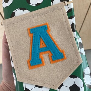 May include: A tan felt banner featuring a large letter "A" in blue with an orange outline. The banner is attached to a gift wrapped in green football-themed paper.