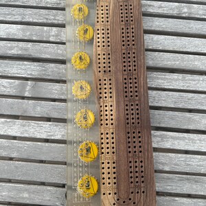 May include: A wooden cribbage board with a clear plastic scoring track. The scoring track has yellow bottle caps with a black triangle warning symbol. The board has a curved track with numbers 1 to 120 and the word "Skunk" printed on it.