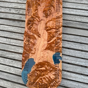 May include: A handcrafted wooden cribbage board with a detailed, textured surface. The board features a natural wood grain with carved details and blue accents. Numerous holes are drilled for peg placement, creating a unique game board.