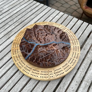 May include: A wooden cribbage board with a detailed, dark brown, topographical map design. A blue river-like line is carved into the map. The board is surrounded by a light brown, circular frame with evenly spaced holes for pegs.