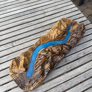 May include: A wooden cribbage board with a river design. The board is made of brown wood with a blue river running through the center. The board has holes for pegs and is set on a wooden table.