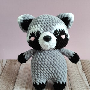 May include: A gray and black crocheted raccoon plush toy with white accents around the eyes and nose. The toy has black eyes and a black nose.