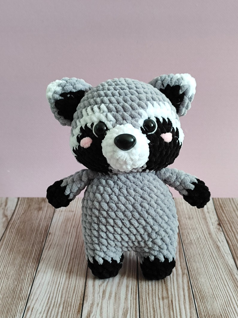 May include: A gray and black crocheted raccoon plush toy with white accents around the eyes and nose. The toy has black eyes and a black nose.