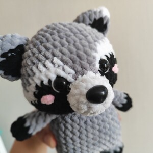 May include: A crocheted raccoon plush toy with gray and white fur, black eyes, a black nose, and pink cheeks.