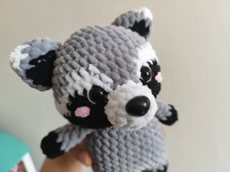 May include: A crocheted raccoon plush toy with gray and white fur, black eyes, a black nose, and pink cheeks.