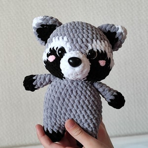 May include: A gray and white crocheted raccoon plush toy with black eyes and a black nose. The toy has pink cheeks and black accents on the ears, paws, and tail.
