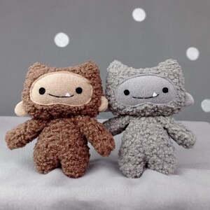 May include: Two plush toys with furry brown and gray bodies and smiling faces. The toys have a simple design with stitched eyes and mouths.