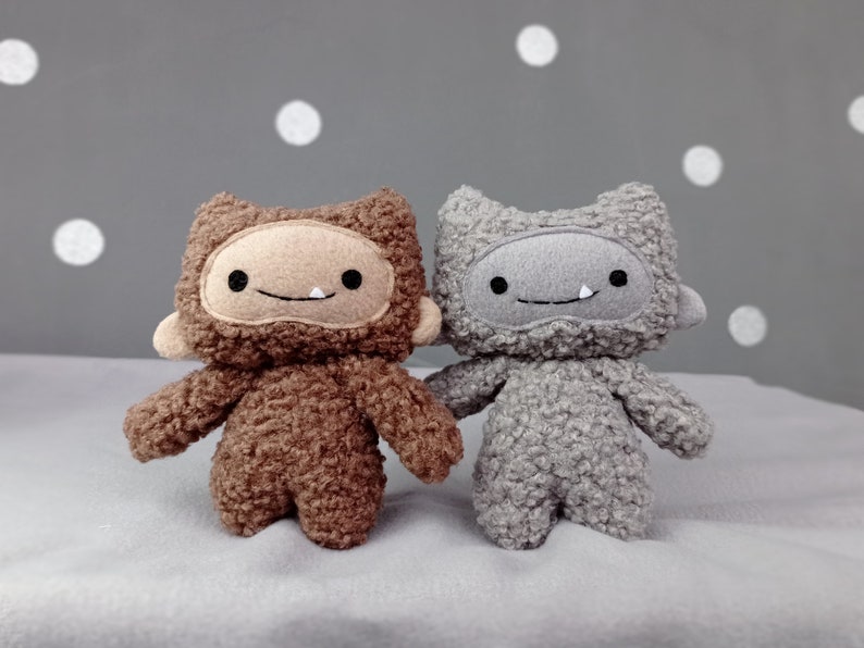 May include: Two plush toys with furry brown and gray bodies and smiling faces. The toys have a simple design with stitched eyes and mouths.