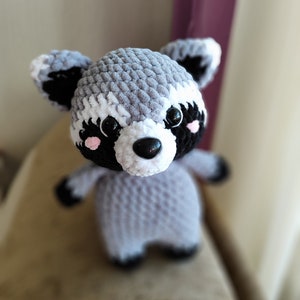 May include: A crocheted raccoon plush toy with black eyes, a black nose, and pink cheeks. The toy is gray and white with black accents.