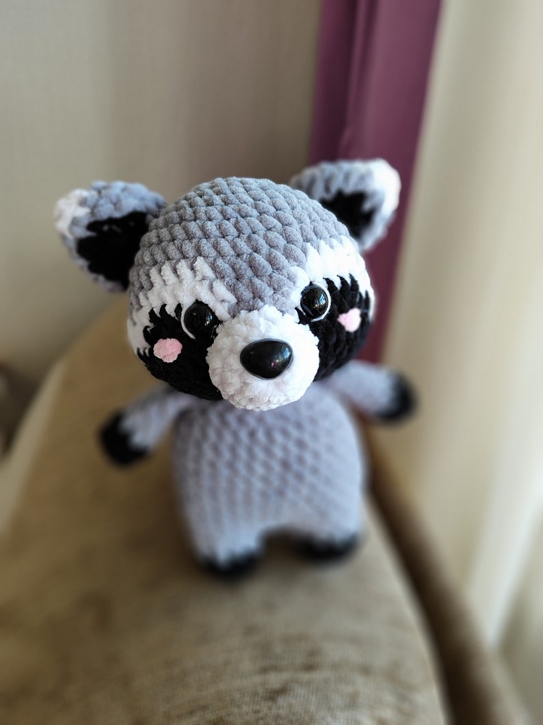 May include: A crocheted raccoon plush toy with black eyes, a black nose, and pink cheeks. The toy is gray and white with black accents.
