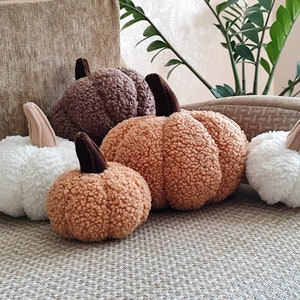 May include: A selection of plush pumpkins in different sizes and colours. The pumpkins are made from a soft, textured material, in shades of white, brown, and orange. Each pumpkin has a brown stem. The pumpkins are arranged on a neutral-toned surface.