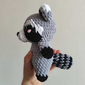 May include: A crocheted raccoon plush toy with gray and black fur, white ears, and pink cheeks. The toy is being held by a hand.
